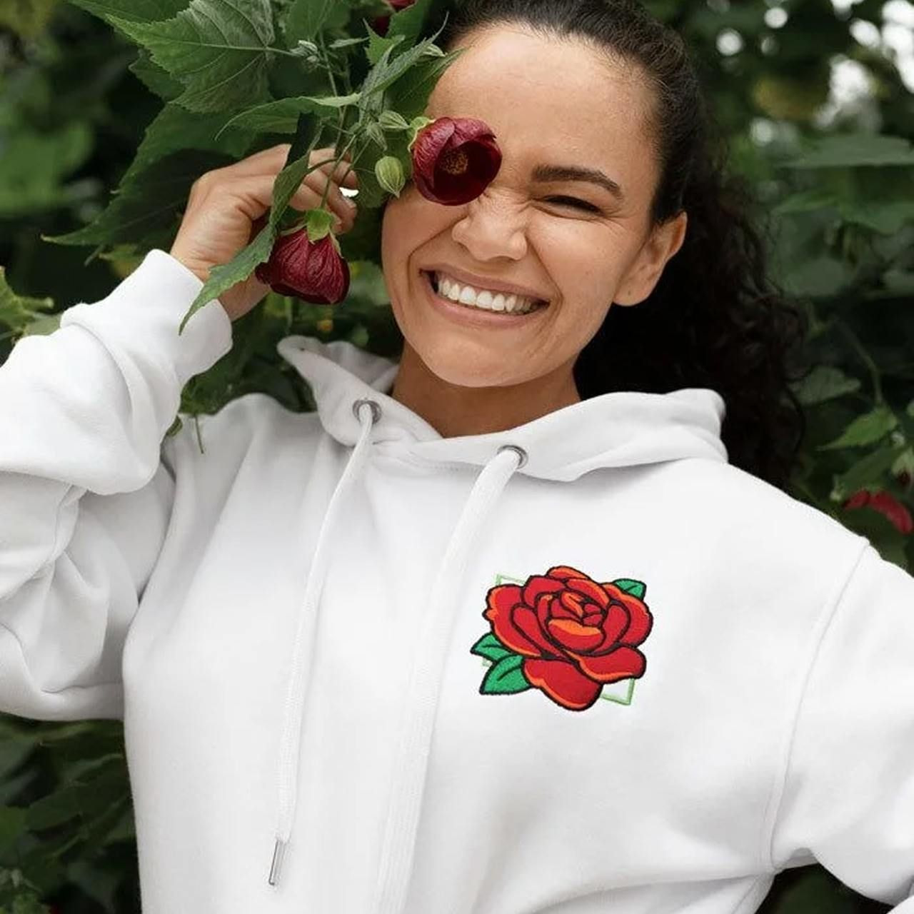 A smiling young woman in a white hoodie with an embroidered rose is holding a wild rose.